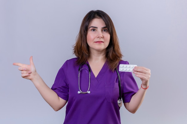 Middle aged woman nurse wearing medical uniform and with stethoscope holding blister with pills with serious face