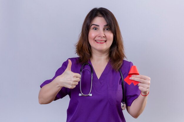 Middle aged woman nurse wearing medical uniform and with stethoscope holding red ribbon a symbol of the fight against aids