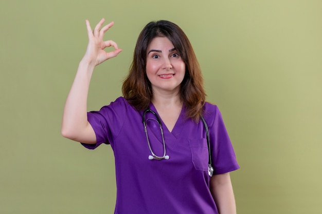 Middle aged woman nurse wearing medical uniform and with stethoscope smiling cheerfully doing ok sign over isolated green wall