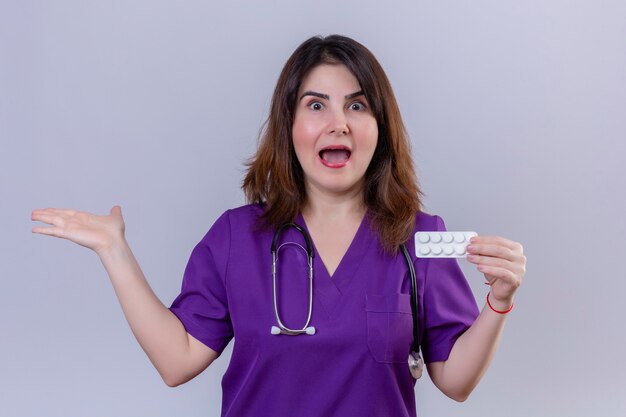 Медсестра средних лет в униформе и со стетоскопом, держащая блистер с таблетками, изумлена и счастлива, указывая ладонью в сторону, стоя на белом фоне