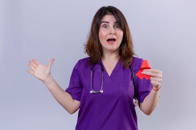 Middle aged woman nurse wearing uniform and with stethoscope holding red ribbon a symbol of the fight against aids amazed and surprised pointing with palm of hand to the side standing over whit