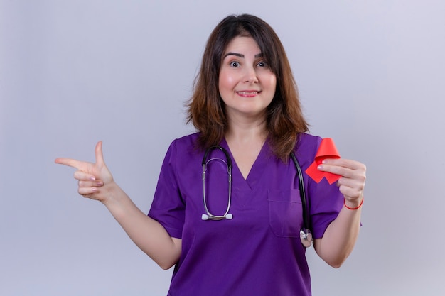 Middle aged woman nurse wearing uniform and with stethoscope holding red ribbon a symbol of the fight against aids smiling friendly pointing to the side with index finger standing over white ba