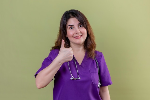 Infermiera donna di mezza età che indossa l'uniforme e con lo stetoscopio che guarda l'obbiettivo sorridente positivo e felice che mostra i pollici in su in piedi sopra il fondo verde