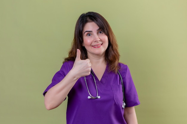 制服を着ている中年女性看護師と聴診器の肯定的で幸せな笑顔で緑の壁に親指を表示