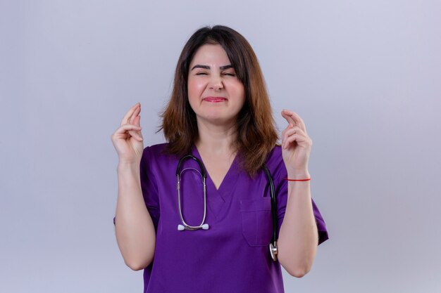 Middle aged woman nurse wearing uniform and with stethoscope with closed eyes raises fingers crossed