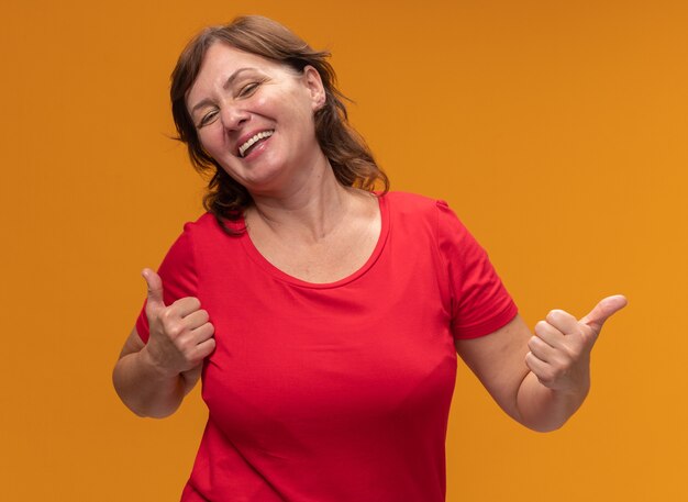 Middle aged woman in red t-shirt  happy and cheerful showing thumbs up standing over orange wall