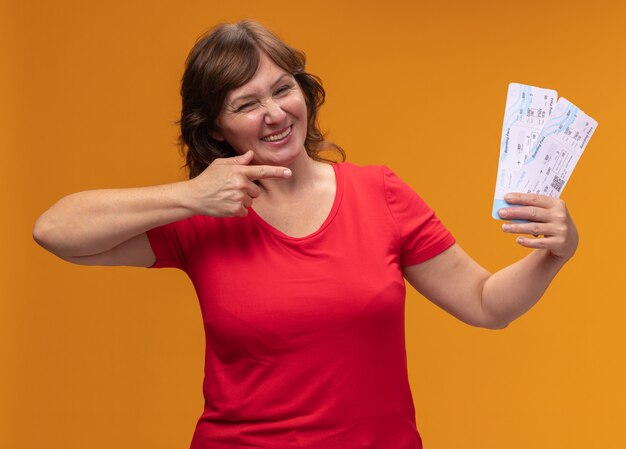 Middle aged woman in red t-shirt holding air tickets pointing with index finger at them happy and positive smiling standing over orange wall