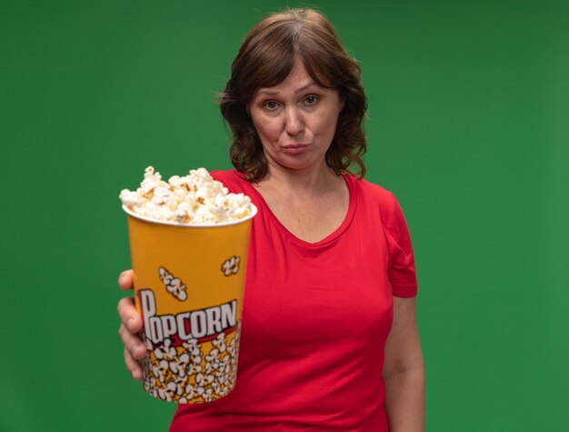 Женщина средних лет в красной футболке, держащая ведро с попкорном с грустным выражением лица, стоит над зеленой стеной