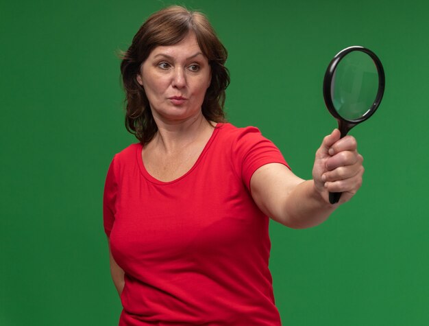 Middle aged woman in red t-shirt holding magnifying glass looking at it with serious face standing over green wall