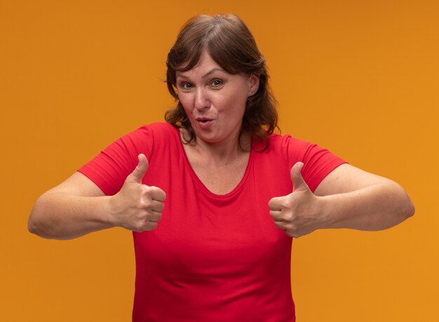 Middle aged woman in red t-shirt  smiling showing thumbs up standing over orange wall