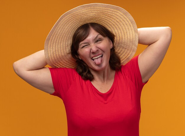 Middle aged woman in red t-shirt and summer hat happy and joyful sticking out tongue standing over orange wall