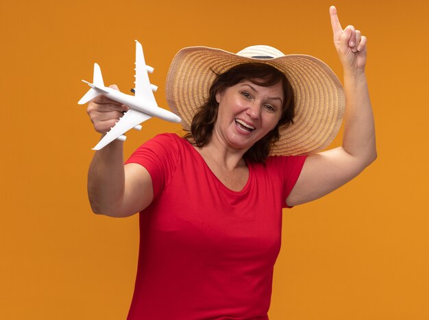 Женщина средних лет в красной футболке и летней шляпе держит игрушечный самолетик, счастливая и взволнованная, указывая пальцем idex вверх, стоя над оранжевой стеной