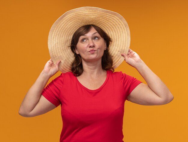 Middle aged woman in red t-shirt and summer hat looking up happy and positive standing over orange wall