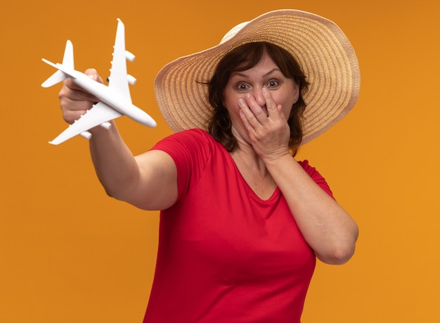 Middle aged woman in red t-shirt and summer hat showing toy airplane  being surprised covering mouth with hand standing over orange wall