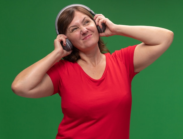 Donna di mezza età in maglietta rossa con le cuffie che ascolta la musica dispiaciuta in piedi sopra la parete verde