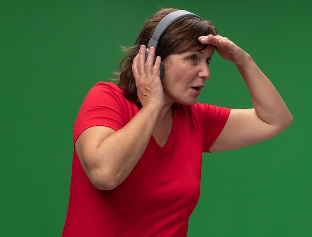 Middle aged woman in red t-shirt with headphones looking far away with hand over head standing over green wall