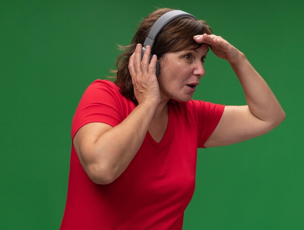 Middle aged woman in red t-shirt with headphones looking far away with hand over head standing over green wall