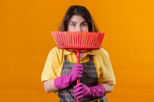 Бесплатное фото Женщина средних лет в фартуке и резиновых перчатках держит швабру, прячась за ней, выглядит удивленной, стоя у оранжевой стены