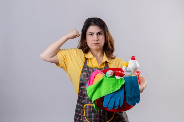 Middle aged woman wearing apron holding bucket with cleaning tools looking displeased with frowning face raising fist standing over white wall