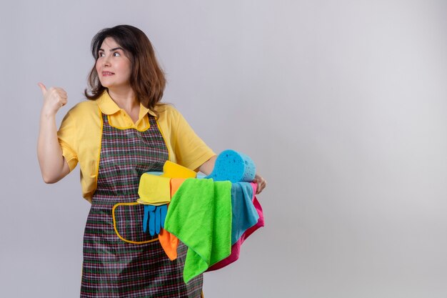 Middle aged woman wearing apron holding bucket with cleaning tools pointing to something behind smiling posotove and happy standing over white wall 2