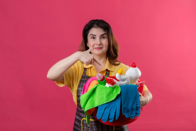 Middle aged woman wearing apron holding bucket with cleaning tools pointing with finger to it smiling standing over pink wall 3