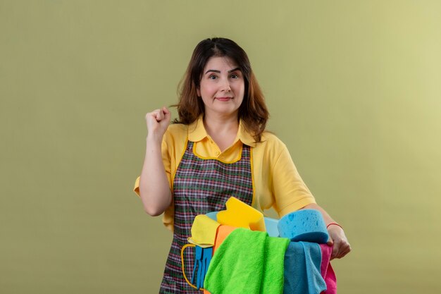 Middle aged woman wearing apron holding bucket with cleaning tools raising fist rejoicing her success smiling looking confident standing over green wall