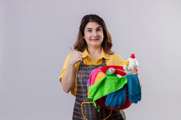 Middle aged woman wearing apron holding bucket with cleaning tools smiling cheerfully positive and happy showing thumbs up standing over white wall 2