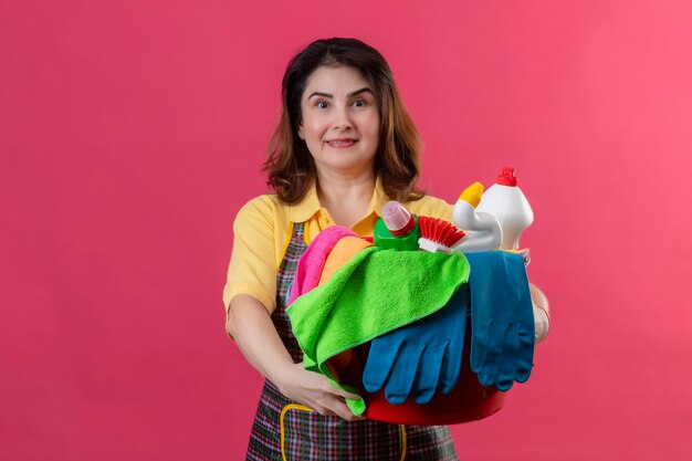 Middle aged woman wearing apron holding bucket with cleaning tools smiling positive and happy standing over pink wall