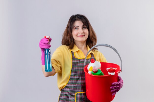 Middle aged woman wearing apron and rubber gloves holding bucket with cleaning tools and cleaning spray positive and happy standing over white wall