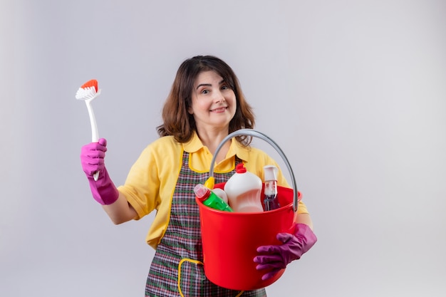 Middle aged woman wearing apron and rubber gloves holding bucket with cleaning tools and scrubbing brush