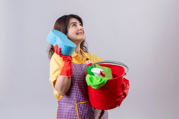 Middle aged woman wearing apron and rubber gloves holding bucket with cleaning tools and sponge