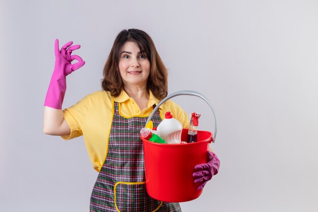 Middle aged woman wearing apron and rubber gloves holding bucket with cleaning tools