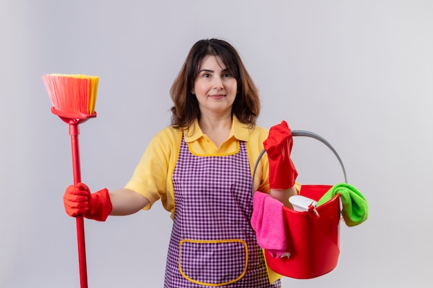Middle aged woman wearing apron and rubber gloves holding bucket with cleaning tools