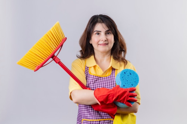 Middle aged woman wearing apron and rubber gloves standing with mops holding sponge smiling cheerfully positive and happy over white wall
