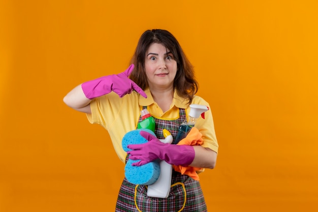 Middle aged woman wearing apron and rubber gloves with cleaning supplies pointing with finger to them smiling friendly positive and happy standing over orange wall 2