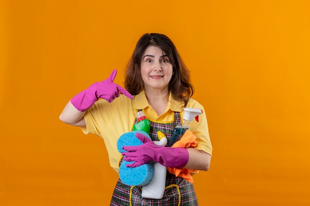 Женщина средних лет в фартуке и рубиновых перчатках с чистящими средствами, указывающими на них пальцем, улыбается дружелюбно и позитивно и счастлива, стоя у оранжевой стены