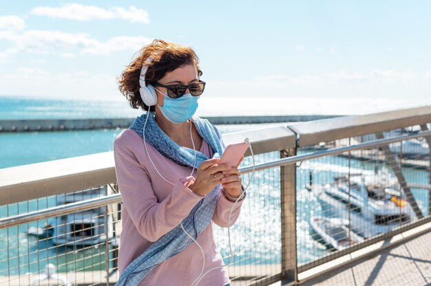 Middle-aged woman wearing a face mask standing on a bridge and listening to music