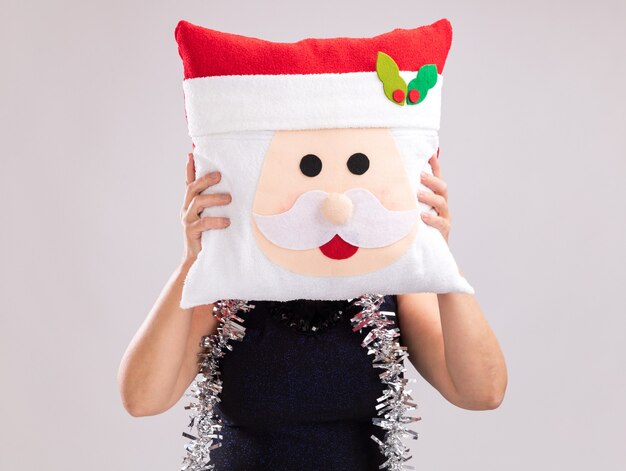 흰색 배경에 고립 된 얼굴 앞에 산타 클로스 베개를 들고 목에 산타 모자와 반짝이 갈 랜드를 입고 중년 여성