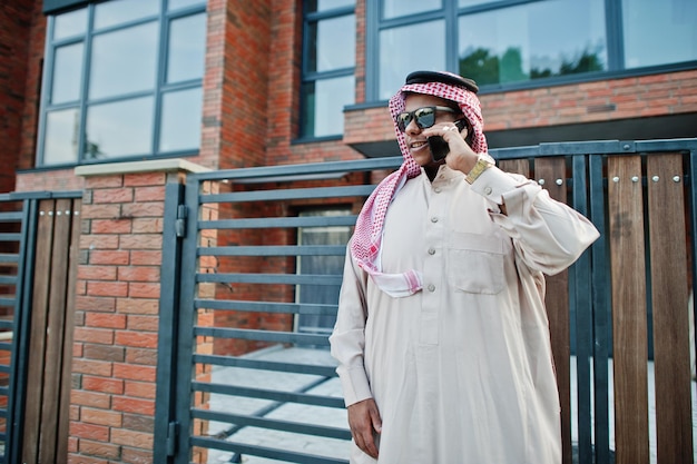 Ближневосточный арабский бизнесмен позирует на улице на фоне современного здания в солнечных очках и разговаривает по мобильному телефону