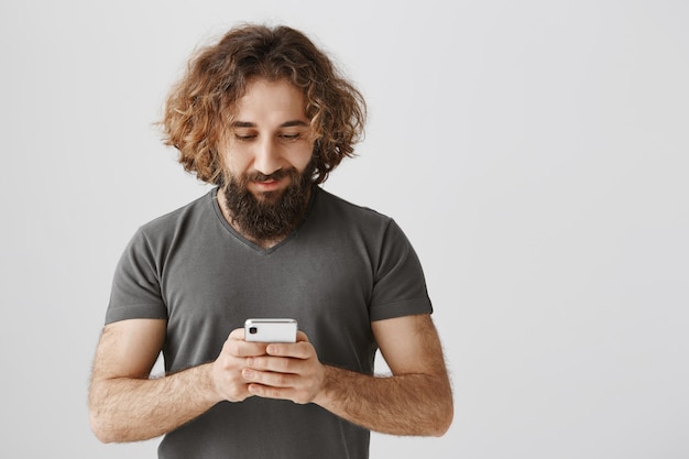 Foto gratuita uomo barbuto mediorientale utilizzando il telefono cellulare, mandare sms e sorridere felice