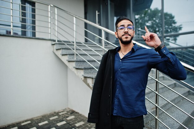 Ближневосточный предприниматель носит черное пальто и очки в синей рубашке на фоне офисного здания, показывает палец вверх