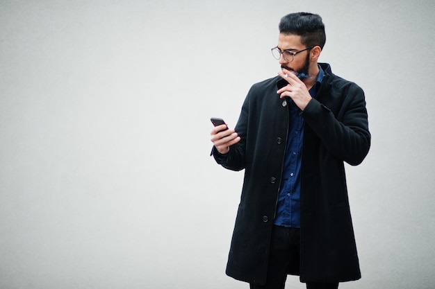 Ближневосточный предприниматель носит черное пальто и очки в синей рубашке, стоя у белой стены, курит сигарету и смотрит в телефон