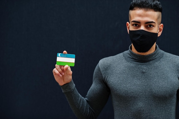 Middle eastern man in gray turtleneck and black face protect mask show uzbekistan flag isolated background