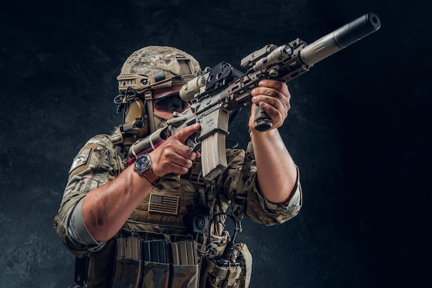 Military man in full equipment with wach on his hand is holding machine gun while posing for photographer over dark background.