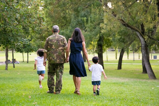 Military man walking in park with his wife and children, kids and parents holding hands. Full length, back view. Family reunion or military father concept