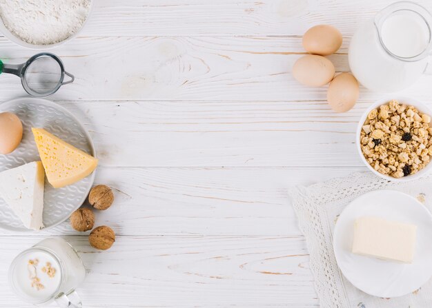 Milk; eggs; bowl of cereals; cheese; flour and walnuts on white wooden table