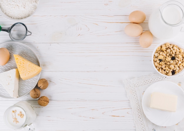 Milk; eggs; bowl of cereals; cheese; flour and walnuts on white wooden table