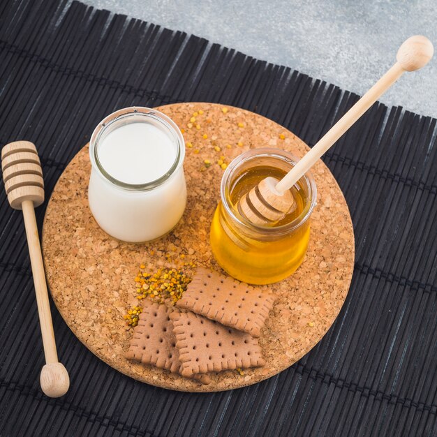 Milk and honey pots with biscuits and bee pollens on cork coaster