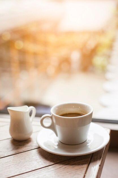 Milk pitcher and coffee cup on wooden table near glass window