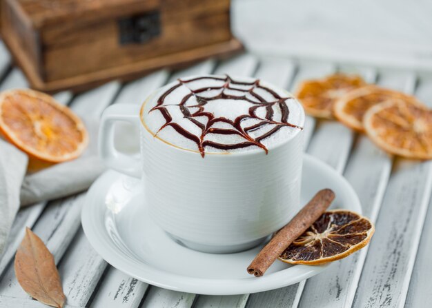 Milky cappuccino with chocolate syrup in white cup with cinnamon and orange slices.